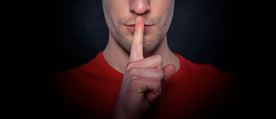 A man wearing a red shirt making a silence gesture with his finger on his lips against a dark background, symbolizing secrecy and confidentiality.