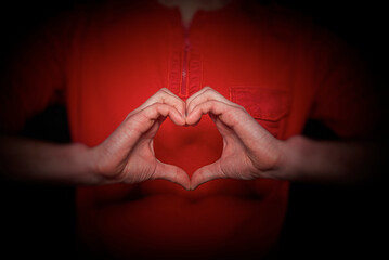 A person wearing a red shirt forming a heart shape with their hands, symbolizing love, care, and positive emotion on a dark background.