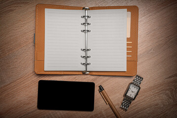 Open personal organizer, a smartphone, a pen, and a wristwatch on a wooden desk.