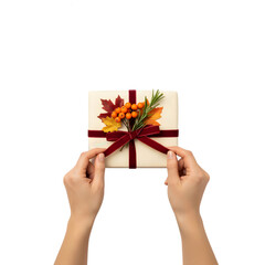 Two hands hold a gift box decorated with autumn leaves and berries. The box is wrapped in white paper with a red ribbon. Transparent background.