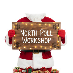Santa Claus in a red suit holds a wooden sign that reads 'NORTH POLE WORKSHOP'. The background is transparent, emphasizing the festive theme.