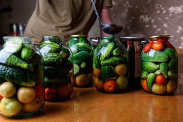 Preservation of season vegetables. A woman pouring the prepared marinade into jars with cucumbers and tomatoes in the kitchen.