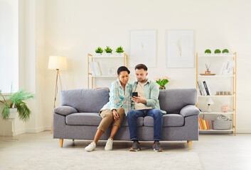 Portrait of young couple sitting on sofa in living room at home and using smartphone together browsing apps. Man showing video or news on mobile phone to his cheerful african american wife.