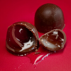 Broken chocolate bonbon with whole cherry and booze syrup spilling on a red background, glossy macro of liquor‑filled candy for party desserts. Photo