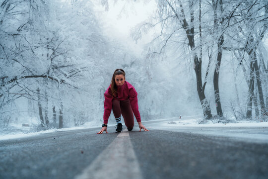 Young woman in athletic wear taking a starting position on a snowy asphalt road surrounded by frost covered trees, concentrating on her winter training and fitness goals - Powered by Adobe