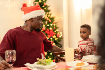 Father and his son enjoying a Christmas dinner together at home. Black man, wearing a Santa hat, sit at a festive table near his young child. Loving bond and togetherness during the winter celebration