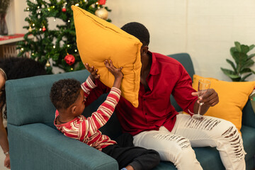 Festive holiday fun with a Black father and son having a playful pillow fight on sofal. Celebrating New Year's Eve or a joyful family Christmas party.