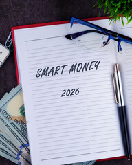 Open notebook displays smart money written boldly on a lined page. Nearby, a pen rests beside stacks of cash and a pair of glasses. A green plant adds a touch of nature to the scene