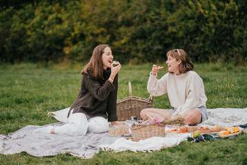 Two women are sitting on a blanket in a park, eating food and laughing. Scene is lighthearted and joyful