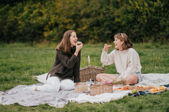 Two women are sitting on a blanket in a park, eating food and laughing. Scene is lighthearted and joyful