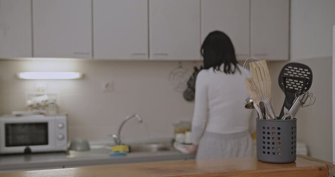 Girl in white top and grey pants washes and wipes dishes and puts them away in her kitchen.