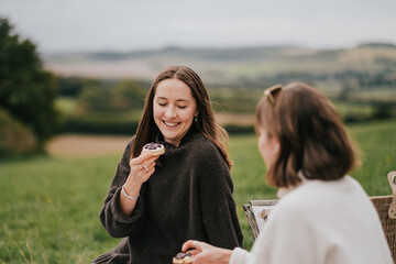 Two women are sitting in a grassy field, eating donuts and smiling. Scene is lighthearted and joyful, as the women are enjoying each other's company and the simple pleasure of eating donuts outdoors