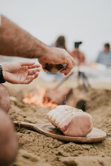 A man sprinkling salt on a piece of meat on a wooden board. The scene is set on a beach, with a fire nearby. The man is surrounded by other people, some of whom are sitting on the sand