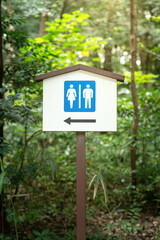 Public toilet direction sign with unisex gender icon and arrow pointing to the left, located in public parkland with greenery tree as background.