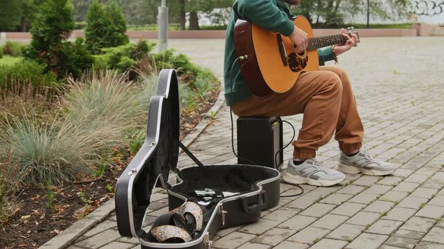 Full shot of unrecognizable male busker sitting on pavement in green park, playing guitar, next to instrument case, while collecting cash donations from passersby and spectators