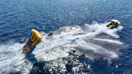 Aerial drone tracking photo of powerboat towing inflatable tubes used for water sports activities in deep blue open ocean island sea