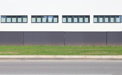 Black concrete fence in front of a modern white industrial building with a rows of windows. Grass and street in front. Background for copy space.