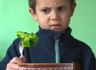 A young boy looks down at a floret of broccoli on a fork with a clear expression of dislike or...