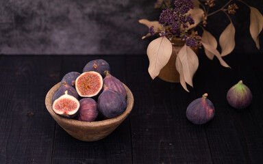 Two sliced figs arranged in a wooden bowl beside whole figs on dark wooden table, moody still life with vase decor in the back