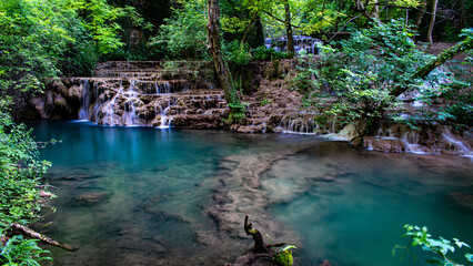 Naklejka premium Long exposure of Krushunski Waterfalls cascading into a turquoise pool surrounded by dense green forest. A serene summer landscape with smooth flowing water and lush vegetation.