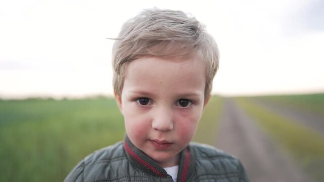Child gazes into camera closeup portrait of boy face in field by dirt road with grass, blond hair, rosy cheek, gray jacket, neutral expression, large eyes, windblown hair, soft sky, distant rural road