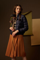Woman in a navy puffer jacket and brown skirt holding dried flowers, standing with a modern atmosphere. © Chuanliang