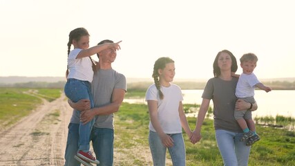 Family walking along lake road at sunset holding hands and carrying child while father smiles and mother steadies toddler near water and grass wearing shirt and jeans together under warm golden light