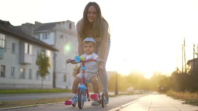 Helping toddler ride tricycle on sunlit street with mother guiding gently while child wears helmet and parent supports learning balance on bike near suburban sidewalk during sunset outdoor family play