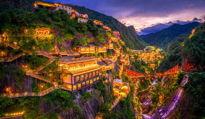 Illuminated ancient village with a red lantern bridge and waterfall built into a steep cliff in a green mountain valley at twilight in Jiangxi.
