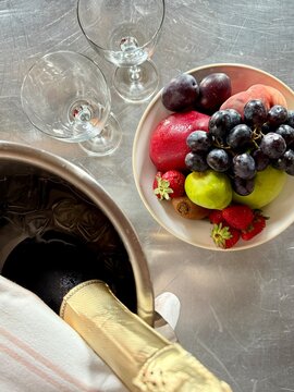Overhead view of a bottle of champagne in an ice bucket with two empty glasses and a bowl of fruit on a table