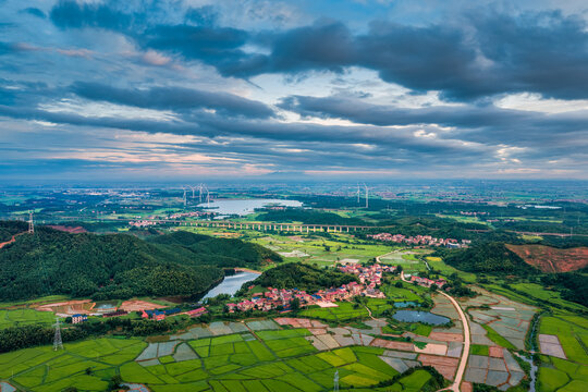 High altitude view of a vast rural landscape with green fields, villages, a lake, and a wind farm under a dramatic sky.