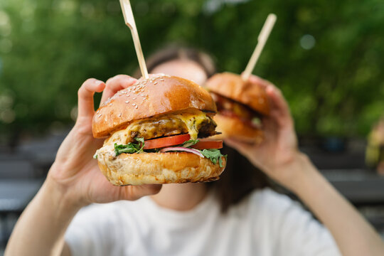 Close-up of a young woman sitting at a garden table holding two juicy cheeseburgers with pickles and salad in front of her face