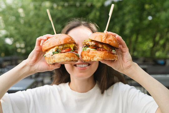 Close-up of a smiling woman sitting at a garden table holding two juicy cheeseburgers with pickles and salad in front of her face