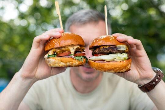 Close-up of a man sitting at a garden table holding two juicy cheeseburger s with pickles and salad in front of his face