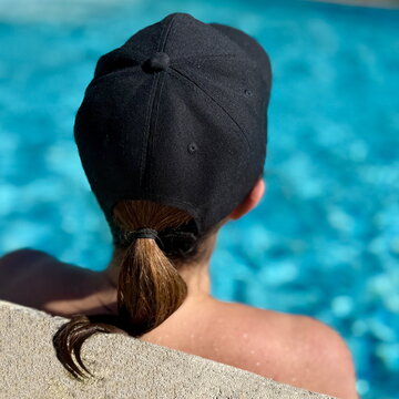 Rear view of a woman in a baseball cap and strapless bikini leaning against the edge of a swimming pool