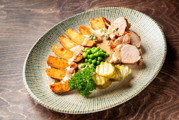 Rustic plated meal with roasted pork tenderloin in sauce, potato wedges, green peas, pickles, and parsley garnish on ceramic plate over wooden table.