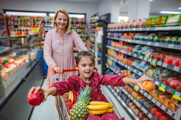 Mother pushing daughter in shopping cart with fruit in grocery store