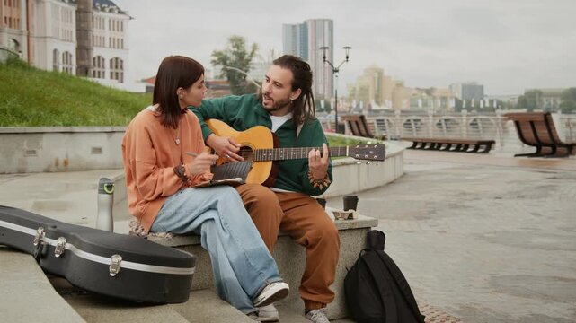 Full shot of young Caucasian busker couple sitting on bench by river on cloudy summer day, composing new song, man playing chords on guitar while girl is writing melody in notebook