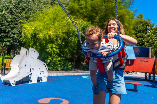 Happy mother pushing baby son on swing in playground