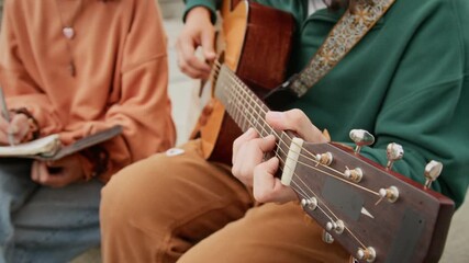 Medium midsection shot of hands of unrecognizable young man playing melody on acoustic guitar outdoors, while anonymous female companion is writing in notebook while composing new song - Powered by Adobe
