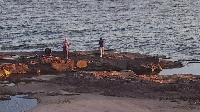 High-angle wide shot with a fixed camera showing two men fishing on the beach, standing on the rocky edge of the R&iacute;o de la Plata shoreline in Montevideo, Uruguay.