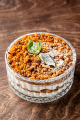 Layered dessert in a glass bowl with crumbled cake, creamy filling, powdered sugar, and mint garnish, served on rustic wood. Top view, close-up, natural light.