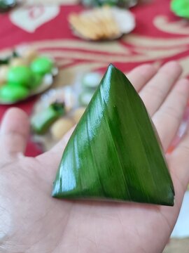 Close-up of a traditional Indonesian snack wrapped in fresh green banana leaf, held by hand. The tringular shape and glossy leaf texture, eco-friendly food packaging 