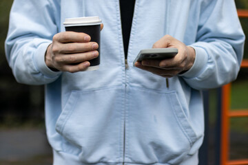 Hands Holding Coffee and Smartphone. Close-up of person's hands in light blue hoodie gripping black takeaway coffee cup and phone, with orange gym bars in background.