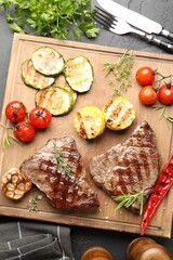 Delicious grilled beef steaks with herbs and vegetables on dark textured table, flat lay