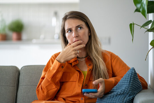 Middle-aged woman deeply thinking providing intense analysis in thoughts while holding phone seated on home sofa. Serious focused female carefully assessing risks and benefits making personal decision