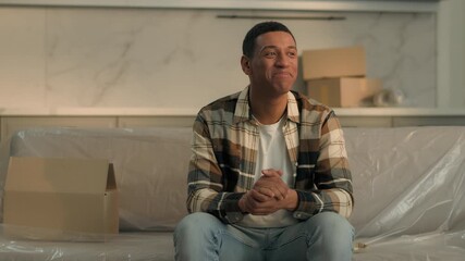 Glad African American man guy male smiling feeling good in new home rental apartment dwelling buying real estate selling property relax on couch near boxes moving relocation buying renovation mortgage