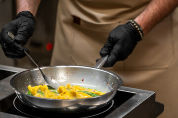 Chef in black gloves cooking creamy pasta with vegetables in stainless steel pan on electric stove, wearing beige apron in professional kitchen setting.