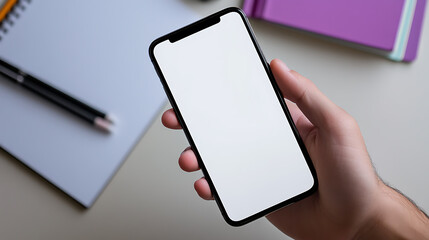 Hand holding a blank screen mobile phone, ready to be customized with your content. Notebook, pen and planner in the background. Workspace ready for creativity.