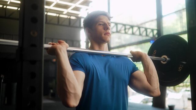 Athletic blonde man in blue t-shirt performing overhead barbell press in spacious modern gym with bright natural lighting and greenery view while demonstrating proper shoulder exercise technique